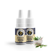 A pair of Blink Ease bottles stand side-by-side against a checkered backdrop. At their feet sits a small white bowl of crushed green herbs and fresh neem leaves, emphasizing the "farm-to-bottle" Ayurvedic philosophy.